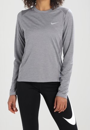 Kvinde iført grå langærmet Nike-trøje og sorte leggings med stort hvidt Nike-logo på låret, står mod en ensfarvet baggrund.
