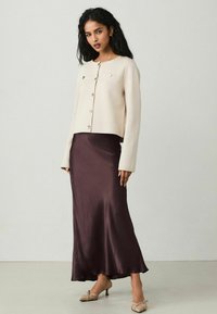 Cardigan cropped beige chiaro con scollatura rotonda e bottoni dorati, abbinato a una gonna midi in satin bordeaux scuro e scarpe beige con tacco.