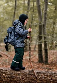 Niño con una chaqueta gris con estampado y pantalones verdes equilibrándose sobre un tronco, sosteniendo un palo. El entorno incluye árboles en un bosque.