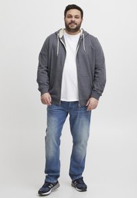 Felpa grigia con zip e cordini bianchi, indossata sopra una maglietta bianca. Abbinata a jeans azzurri chiari e scarpe da corsa nere.