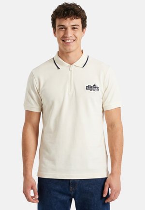 Jeune homme souriant portant un polo crème à manches courtes avec une fermeture éclair et un logo sur la poitrine, associé à un jean bleu.