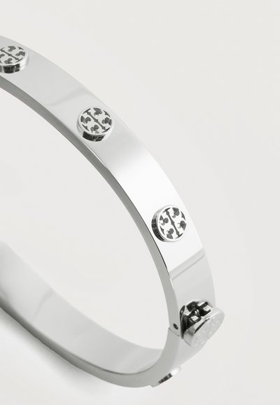 Ασημένιο μεταλλικό βραχιόλι με ομοιόμορφα τοποθετημένα στρογγυλά ανάγλυφα μοτίβα που θυμίζουν τριφύλλι γύρω από τη ζώνη και ασφαλές κούμπωμα κλεισίματος.