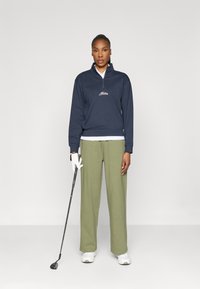 Námořnický zipový pulovr, bílá značka a zelené široké kalhoty. Model drží golfovou hůl a nosí bílé tenisky a rukavici. Minimalistické pozadí.