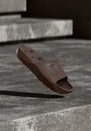 Hnedá textúrovaná slide sandal s logom plávajúcim nad betónovým povrchom a schodmi v pozadí.