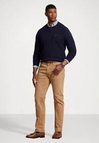 Πλεκτό πουλόβερ ναυτικού τύπου φορεμένο πάνω από ένα ανοιχτό μπλε πουκάμισο με κουμπιά, συνδυασμένο με καφέ παντελόνι chinos και καφέ μοκασίνια. Ρολόι στον καρπό, ουδέτερο φόντο.