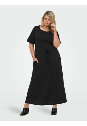 Kvinde iført en sort maxi-kjole med korte ærmer, taljebånd og lommer, kombineret med sorte hælede sko, stående på en ensfarvet baggrund.