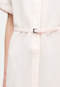 Κοντινό πλάνο ενός ατόμου που φοράει ανοιχτό ροζ φόρεμα με κοντά μανίκια, με στενή ζώνη και μικρή μεταλλική αγκράφα στη μέση.