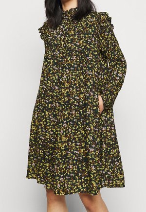 Femme portant une robe fleurie foncée longueur genou avec manches longues, col haut et épaules à volants, main dans la poche latérale.