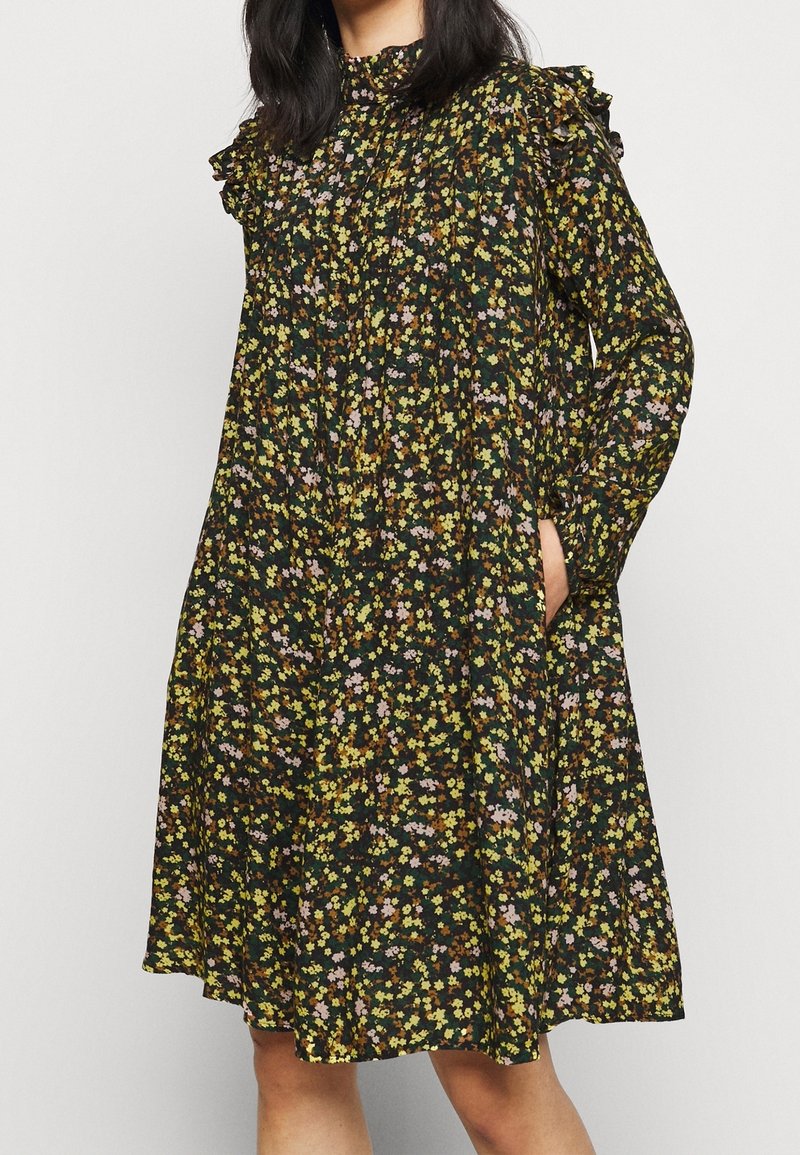 Femme portant une robe fleurie foncée longueur genou avec manches longues, col haut et épaules à volants, main dans la poche latérale.