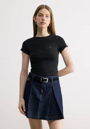 Fiatal nő fekete, testhezálló rövid ujjú felsőt és sötétkék farmer szoknyát fekete övvel viselve, egyszerű háttér előtt áll.