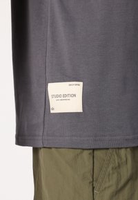 Gray t-shirt featuring a cream label with "STUDIO EDITION" text. Made of soft fabric with a classic fit and short sleeves.