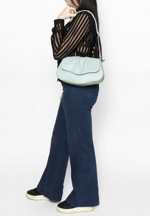 Borsa a spalla in pelle verde menta con parte superiore curva, superficie texturizzata e una sola tracolla lunga, abbinata a pantaloni blu scuro svasati e scarpe nere.