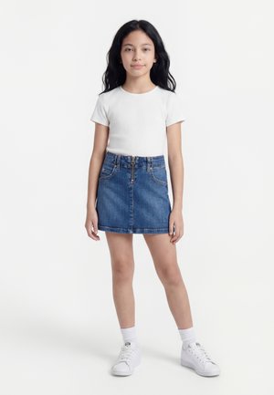 Fată cu păr lung și negru poartă un top alb cu mâneci scurte, fustă din denim, șosete albe și adidași albi, stând în picioare pe un fundal simplu.