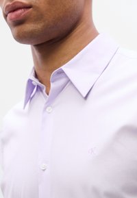 Ανοιχτό μωβ πουκάμισο με κλασικό γιακά, δύο κουμπιά και μικρό κεντητό λογότυπο στο αριστερό στήθος. Ομαλή υφή υφάσματος.