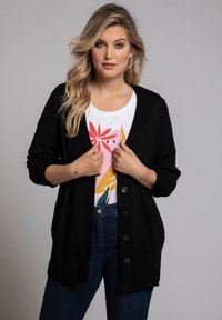 Schwarzer Cardigan mit V-Ausschnitt, durchgehender Knopfleiste und gerippten Bündchen. Tragen über einem weißen T-Shirt mit Grafik und dunklen Jeans.