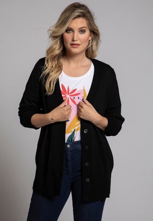 Cardigan noir avec un design en V, fermeture à boutons sur le devant et poignets côtelés. Porté sur un t-Shirt graphique blanc et un jean sombre.