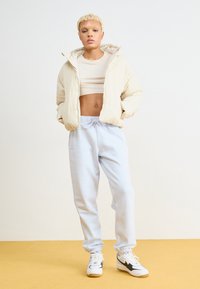 Casaco puffer bege com capuz, top cropped branco, calças de fato de treino azul claro e sapatilhas brancas com detalhes pretos. Tecidos suaves e corte descontraído.