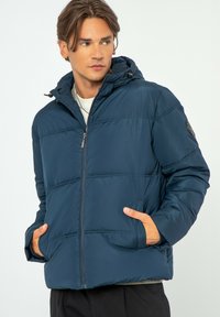 Marineblauwe gewatteerde jas met een ritssluiting aan de voorkant, verstelbare capuchon en twee voorzakken. Kenmerkt zich door horizontale stiksels en een logo-embleem op de mouw.