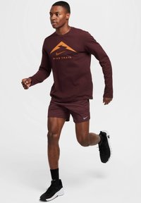 Maroon pitkähihaine paita, jossa on oranssi Nike Trail -grafiikka, yhdistettynä marooniin shortsiin ja mustiin juoksukenkiin, joissa on valkoiset pohjat.