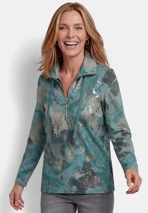 Frau lächelt und trägt einen langärmligen Pullover mit türkis-grauem Muster, halbem Reißverschluss und Kordelkragen, kombiniert mit grauer Hose.