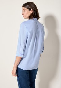 Blusa azzurro chiaro con design casual, caratterizzata da un colletto con bottoni, maniche arrotolabili e dettaglio testuale sulla schiena. Tessuto di trama liscia.