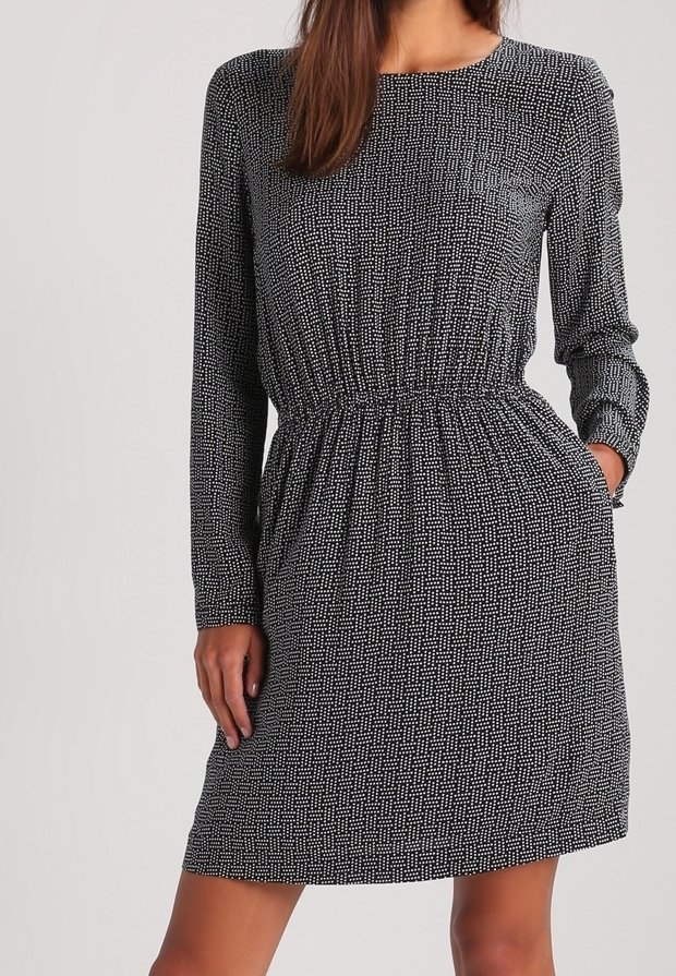Femme portant une robe noire à manches longues avec un motif à pois blancs et une taille cintrée, debout avec une main dans la poche sur un fond uni.