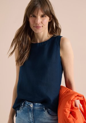 Donna con capelli castani lunghi che indossa una canotta blu scuro e jeans blu, tenendo una giacca trapuntata arancione su uno sfondo beige.