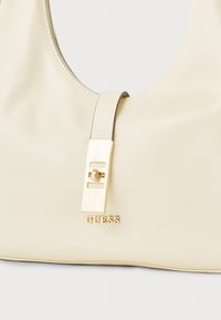 Crèmekleurige leren handtas met een zilverkleurige ritssluiting, een gouden sluiting en het gegraveerde "GUESS"-logo aan de voorkant. Gladde textuur en gebogen vorm.