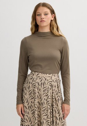 Mujer con cabello largo y rubio, vistiendo una blusa de manga larga verde oliva y una falda beige con un patrón de hojas marrón oscuro, de pie contra un fondo blanco.