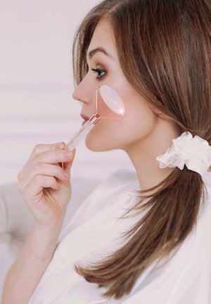 Rouleau facial en or rose maintenu contre une joue, avec une poignée transparente et une pierre ovale, sur fond rose clair.