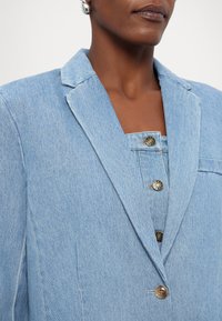 Gros plan d'une personne portant un blazer en denim bleu clair sur un vêtement boutonné assorti, avec des boucles d'oreilles créoles argentées visibles.