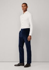 Bijela košulja na dugme s smešnim dugmadi, uparena s tamnoplavim korzetskim hlačama. Model nosi smeđe kožne cipele; jednostavna pozadina.