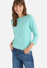 Top largo de color turquesa claro con cuello redondo, diseño ajustado y textura suave. Combinado con jeans azules, con las manos en los bolsillos.