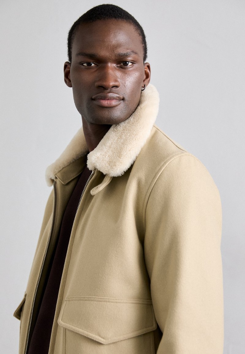 Beige wollen Blend jas met een nepbont kraag, ritsluiting en twee voorkanten zakken. Glad textuur met een getailleerde pasvorm.