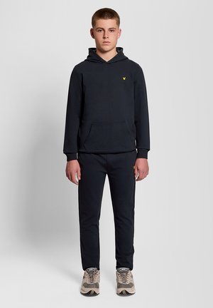 Tienerjongen staand, gekleed in een zwarte hoodie met een geel logo, bijpassende zwarte broek en beige New Balance sneakers, tegen een effen lichte achtergrond.