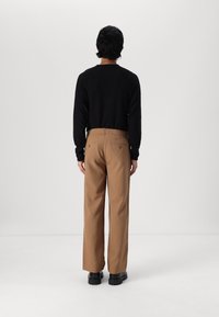 Man som står vänd bort, iklädd en svart långärmad tröja, bruna byxor med bakfickor och svarta kraftiga skor mot en enfärgad bakgrund.