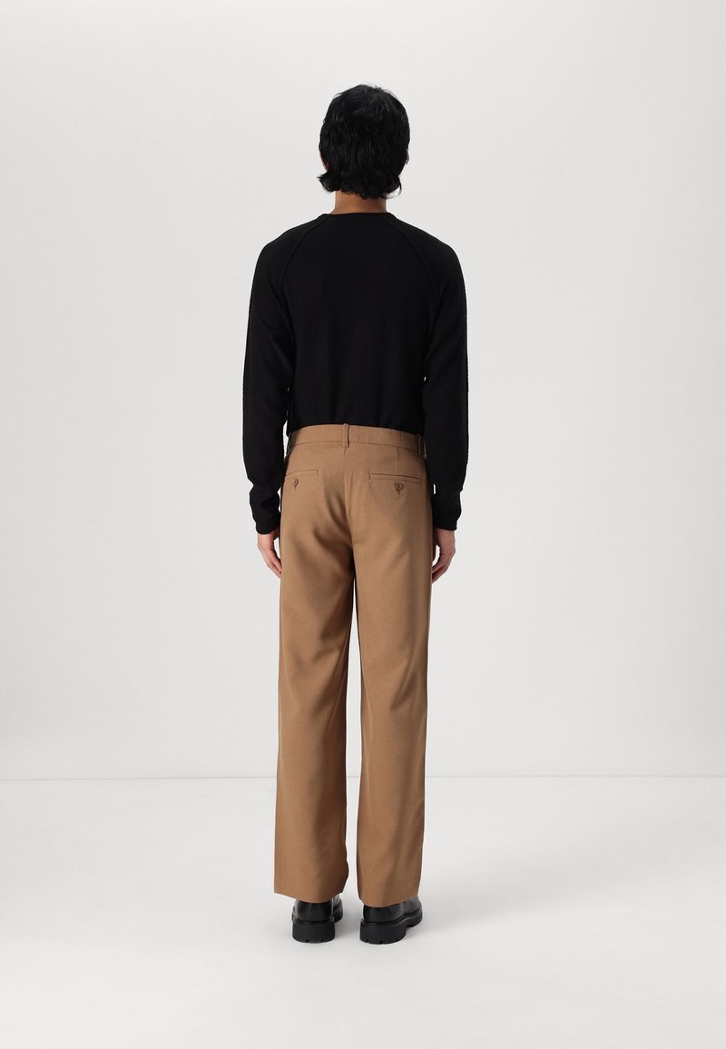 Man som står vänd bort, iklädd en svart långärmad tröja, bruna byxor med bakfickor och svarta kraftiga skor mot en enfärgad bakgrund.