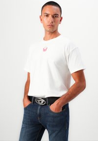 Tricou alb din bumbac cu un mic grafic roșu pe piept, purtat cu blugi negri din denim și o curea neagră cu o cataramă metalică argintie.