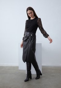 Crna majica s dugim rukavima i prozirnim rukavima s točkicama, uparena s crnom kožnom suknjom na preklop s bočnim gumbom, crne čizme do koljena.