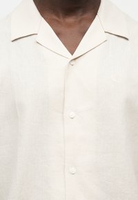 Άτομο με σκούρο δέρμα που φοράει μπεζ πουκάμισο με κουμπιά και γιακά τύπου camp, εμφανίζοντας τρία κουμπιά και διακριτή υφή υφάσματος.