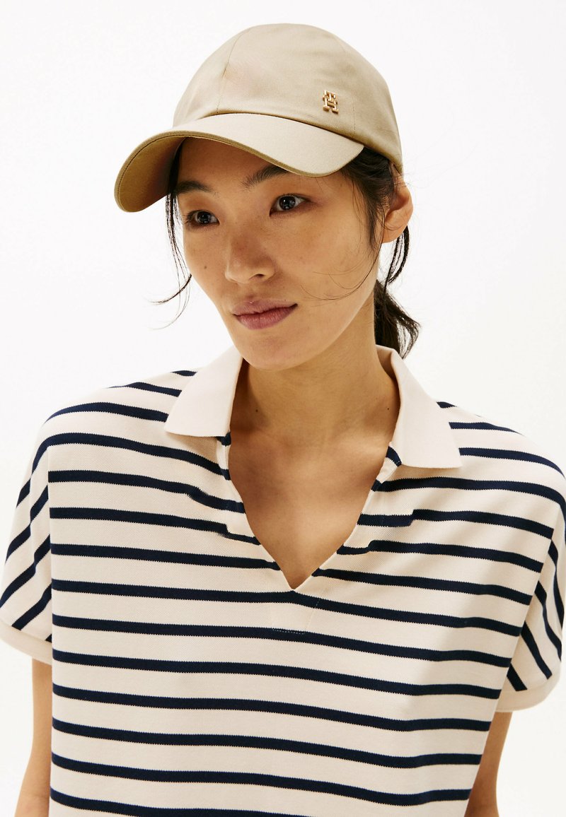 Cappello beige con visiera curvata e piccolo dettaglio del logo. Indossato con una polo a righe con strisce orizzontali blu navy e crema.