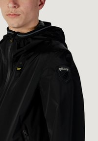 Giacca impermeabile nera con cappuccio, dotata di zip frontale, tasche laterali e un patch con logo sulla manica. Texture liscia e lucida.