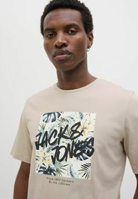 Camiseta de algodón beige con un gran estampado gráfico de hojas y flores tropicales. Texto "JACK & JONES" en negro, exhibido de manera prominente.