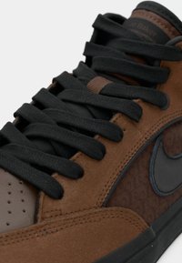 Nahaufnahme eines braunen und schwarzen Sneakers mit schwarzen Schnürsenkeln, Wildleder- und Lederdetails sowie einem sichtbaren Markenlogo an der Seite und der Zunge.