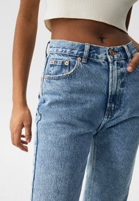Sección media de una persona que lleva jeans de mezclilla de tiro alto en un tono azul claro y una blusa blanca sin mangas y corta frente a un fondo liso.