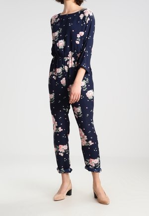 Femme portant une combinaison bleu marine à motif floral avec des poignets retroussés et des chaussures à talons carrés beige, debout devant un fond clair uni.