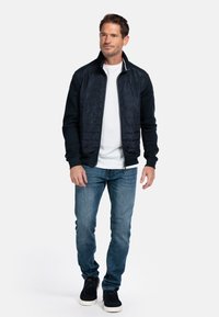 Zwarte, met rits gesloten jack met een gewatteerde voorkant en effen mouwen, gedragen over een wit shirt. Gecombineerd met blauwe jeans en zwarte sneakers.