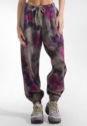Frau trägt lockere Batik-Jogginghosen in Lila-, Pink- und Grautönen, mit elastischen Bündchen und Kordelzug am Bund sowie graue Sneaker.