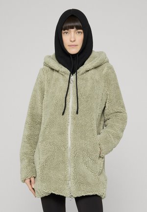 Frau trägt eine hellgrüne, flauschige Jacke mit Reißverschluss und Kapuze sowie eine schwarze Strickhaube, steht vor einem schlichten Hintergrund.