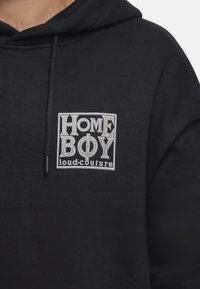 Felpa nera con cappuccio caratterizzata da un'etichetta grafica frontale in testo argento: "HOME BOY loud-couture". Il tessuto appare morbido con una finitura testurizzata.